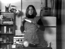 Video still of Martha Rosler in a kitchen holding up a knife, from Semiotics of the Kitchen, 1975.